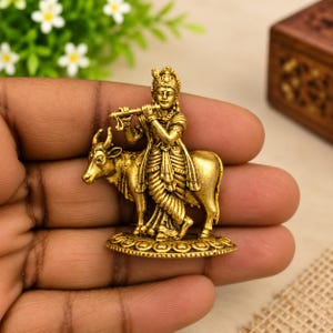 May include: A small, gold-toned metal figurine depicting a standing figure playing a flute while leaning against a cow. The sculpture features intricate detailing on the clothing and the animal, all set upon a circular, textured base.