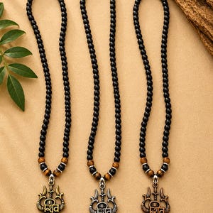 May include: Three necklaces featuring black spherical beads, brown wood-toned accent beads, and metallic trident pendants. Each pendant displays the Devanagari script for Shiv and a central horizontal line symbol, finished in gold, silver, and copper tones.