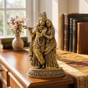 May include: A gold-toned resin statue depicting two figures standing together on an ornate oval base. One figure holds a flute to their lips while wearing a crown and traditional garments, while the other figure stands closely beside them in a long dress.