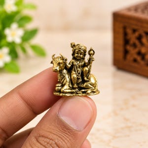 Könnte beinhalten: Eine kleine Messingfigur mit Antik-Finish, die eine Figur auf einer Kuh sitzend darstellt. Die Figur trägt eine Krone und hält einen Stab in der Hand. Die Kuh befindet sich in einer ruhenden Position. Das Stück weist detaillierte Reliefs auf.