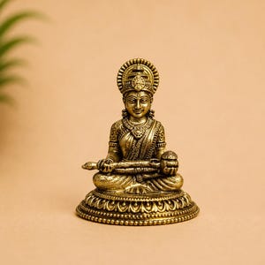 May include: A metallic gold-toned figurine of a seated deity with an ornate headdress, wearing traditional clothing with detailed patterns. The figure sits in a cross-legged position on a circular, decorative base, holding a staff and a small bowl.
