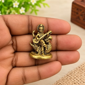 Könnte beinhalten: Eine kleine, messingfarbene Metallfigur im Antik-Stil, die eine sitzende Gottheit darstellt, welche ein Saiteninstrument, eine Veena, spielt und auf einem Schwan ruht. Die detaillierte Skulptur verfügt über einen runden Sockel und ein Metallfinish.