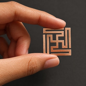 Peut inclure: Un petit pendentif métallique de couleur cuivre, de forme carrée, présentant un motif géométrique en forme de labyrinthe avec une découpe centrale en croix. L'objet a une finition mate et plate, tenu entre les doigts pour montrer sa taille.