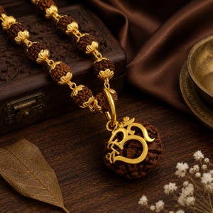Op de afbeelding: Een ketting met bruine, getextureerde rudraksha-kralen gescheiden door gepolijste goudkleurige metalen kapjes. De hanger bestaat uit een grote, ronde rudraksha-kraal versierd met een opvallend, glanzend goudkleurig Om-symbool aan een metalen ring.