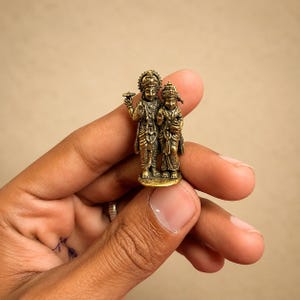 May include: A small, detailed brass figurine depicting two standing figures adorned in traditional attire and ornate headpieces. The metallic sculpture features intricate carvings on the clothing and jewellery, with one figure holding a small circular object.
