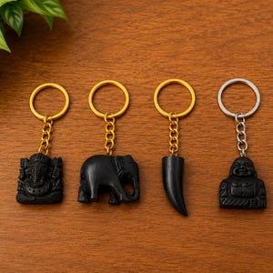 May include: Four keychains featuring black carved charms: a Ganesha figure, an elephant, a curved tusk, and a laughing Buddha. The first three charms hang from gold-toned metal rings and chains, while the fourth hangs from a silver-toned ring.