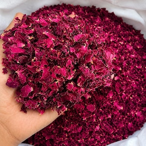 May include: A large quantity of dried, deep magenta rose petals with a textured, crinkled appearance. The petals are gathered in a heap, with a portion held up to display the vibrant, dark pink colour and the dry, brittle nature of the floral material.