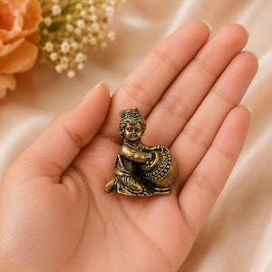 May include: A small, antique-style brass-colored metal figurine depicting a seated child figure with curly hair, wearing simple garments and jewelry, reaching toward a round, textured pot or container held in front of the body.