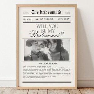 Può includere: Una stampa incorniciata progettata come una prima pagina di giornale, con il titolo "The Bridesmaid". Presenta una foto in bianco e nero di due donne e il testo "Will You Be My Bridesmaid?" La stampa è in una cornice di legno chiaro.