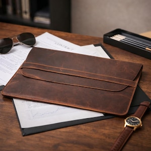 Könnte beinhalten: Eine braune Laptop-Hülle aus Leder mit Klappenverschluss, die auf einem Schreibtisch mit Dokumenten und einer Uhr liegt. Die Hülle hat eine strukturierte Oberfläche und ein genähtes Design. Sonnenbrille und Bleistifte sind ebenfalls sichtbar.