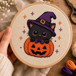 May include: A cross-stitch embroidery project in a round wooden hoop featuring a black cat wearing a purple witch hat and a purple bow tie, sitting inside an orange jack-o'-lantern pumpkin, surrounded by small orange and purple star icons.
