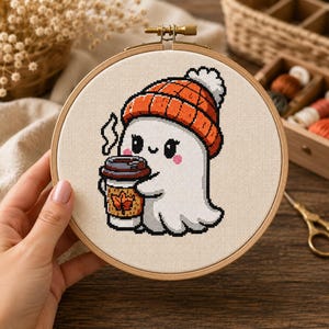 Pode incluir: Um bastidor de bordado circular de madeira contendo um padrão de ponto cruz de um fantasma branco usando um gorro de tricô laranja com um pompom branco. O fantasma segura um copo de café com tampa castanha e uma manga amarela com uma folha.