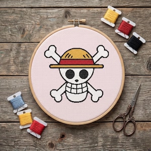 May include: A cross-stitch embroidery hoop featuring a skull and crossbones design with a straw hat. The design is stitched on pink fabric and framed in a wooden hoop. Spools of thread in various colors and a pair of scissors are also visible.
