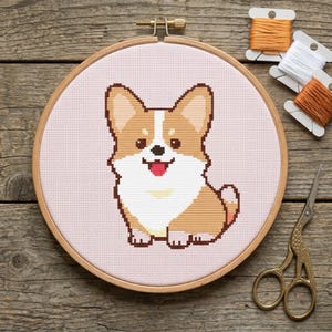 Puede incluir: Un bordado de punto de cruz de un perro corgi sonriente en un aro de madera. El corgi es marrón, blanco y canela, con un fondo rosa. Bobinas de hilo naranja y blanco y tijeras doradas están junto al aro sobre una superficie de madera.