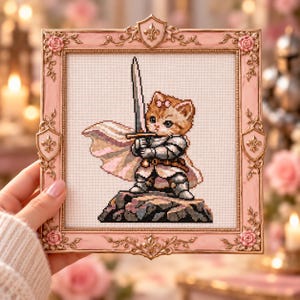 Peut inclure: Une broderie au point de croix représentant un chaton roux portant une armure en plaques argentée et une cape flottante, tenant une longue épée sur un rocher. L'œuvre est encadrée dans un cadre carré, orné, rose clair avec des détails dorés.