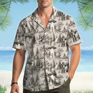 May include: A short-sleeved button-down shirt in a light beige color featuring an all-over black and white graphic print of Bigfoot, extraterrestrial beings, flying saucers, pine trees, and camping tents in a forest setting.