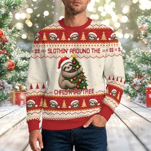 Könnte beinhalten: Ein cremefarbenes Langarm-Sweatshirt mit einem rot-weißen Weihnachtsmuster aus Faultiergesichtern und Tannenbäumen. Die Grafik in der Mitte zeigt ein Faultier mit Weihnachtsmütze, das einen geschmückten Baum umarmt, mit dem Text SLOTHIN' AROUND THE CHRISTMAS TREE.