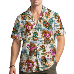 Può includere: Camicia a maniche corte con bottoni con un motivo ripetuto di Bigfoot che trasporta un fenicottero rosa, maschere tiki, ananas e fogliame tropicale. La camicia è principalmente beige con accenti blu, verdi, marroni e rosa.