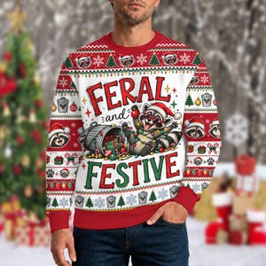Peut inclure: Un sweat-shirt blanc à manches longues avec un motif de vacances rouge et vert représentant un raton laveur portant un bonnet de Père Noël et des lunettes, emmêlé dans des guirlandes lumineuses près d'une poubelle, avec le texte FERAL and FESTIVE.