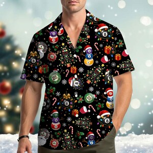 Op de afbeelding: Een zwart overhemd met korte mouwen en knoopsluiting, voorzien van een herhalend kerstpatroon met biljartballen, sneeuwpoppen gemaakt van biljartballen, kerstbomen, cadeautjes, zuurstokken en sneeuwvlokken.