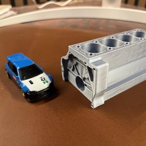 May include: A blue and white toy car with the number "42" and a green "R" on the front, next to a silver engine block model. The car has black tyres and a black front bumper. The engine block has four cylinders.