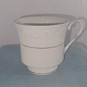 May include: A white teacup with a handle, featuring a decorative floral pattern near the rim and a thin silver band. The cup has a flared top and a pedestal base, suitable for serving hot beverages.