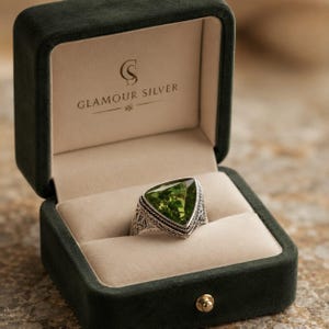 Peut inclure: Bague en argent ornée d'une grande pierre verte triangulaire. La bague est sertie dans un anneau orné et détaillé, présentée dans un écrin en velours vert foncé. Le texte "GLAMOUR SILVER" est visible.