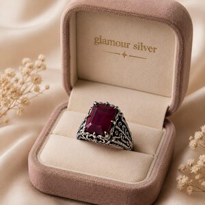 Op de afbeelding: Een zilveren ring met een grote, rechthoekige, dieprode edelsteen. De ring heeft een sierlijk filigraanontwerp en wordt gepresenteerd in een roze fluwelen ringdoos. De doos is bedrukt met de tekst "glamour silver".