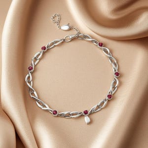 Può includere: Un bracciale a catena color argento caratterizzato da una serie di maglie metalliche intrecciate alternate a piccole pietre preziose rotonde di colore rosso scuro. Il bracciale include una sottile catenella di estensione con un ciondolo a foglia.