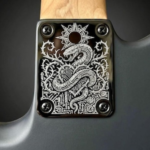 May include: Close-up of a black guitar neck plate with a detailed white engraved design. The design features a snake coiled around a circuit board, with a sunburst and thorns. The plate is secured with four screws.