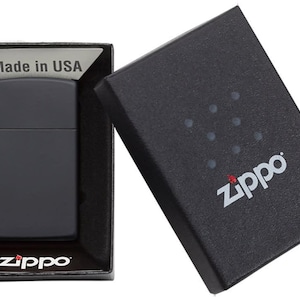 以下が含まれることがあります： 黒色のマットなZippoライターが黒い箱に入っています。箱は開いており、ライターと「Made in USA」の文字が見えます。Zippoのロゴが箱とライターに表示されています。