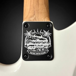 May include: Close-up of an electric guitar's neck joint. A black plate with a white graphic of a sunset, palm trees, and the text "ELECTRIC GUITAR PICKUPS" is attached to the white body and wooden neck.