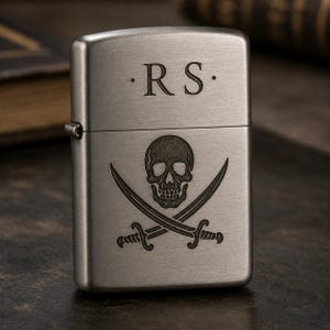 以下が含まれることがあります： 上部に「RS」のイニシャルが刻まれた、つや消しメタルのライター。イニシャルの下には、スカルと交差した骨のデザインがあります。ライターは閉じており、左側にヒンジが付いています。