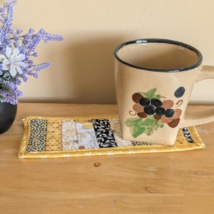 Peut inclure: Un dessous de verre rectangulaire en tissu avec un motif patchwork composé de bandes jaunes, noires, blanches et grises, incluant des imprimés alvéolés et floraux. Une tasse en céramique beige ornée de raisins et de feuilles vertes est posée dessus.