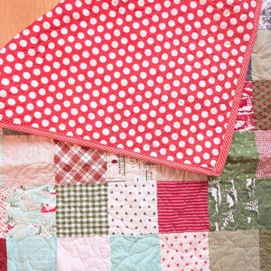 May include: A patchwork quilt featuring a variety of square fabric blocks in shades of red, green, white, and light blue. The quilt is folded over to reveal a bright red backing fabric with a repeating pattern of small white polka dots.
