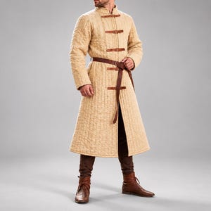 Peut inclure: Un long manteau matelassé beige avec des coutures verticales, un col montant et une fermeture frontale composée de quatre lanières en cuir marron avec boucles. Une ceinture en cuir marron assortie est nouée à la taille sur un pantalon marron.