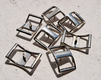 May include: A group of eight silver-toned metal buckles, each with a curved shape and a single hole in the center. The buckles are arranged in a random pattern on a white surface.
