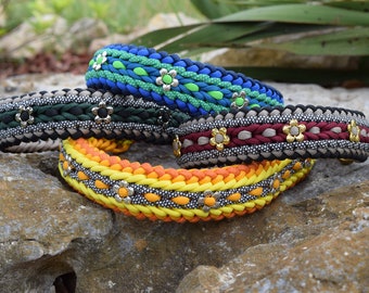 May include: Four colourful dog collars with silver flower accents. The collars are made of braided paracord and feature different colour combinations, including blue, green, black, grey, red, and yellow.