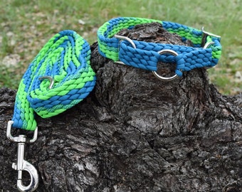 Puede incluir: Un juego de collar y correa trenzados para perros. El collar y la correa son azules y verdes, con herrajes metálicos plateados. La correa está enrollada junto al collar. El conjunto se muestra sobre una superficie texturizada de color marrón oscuro.