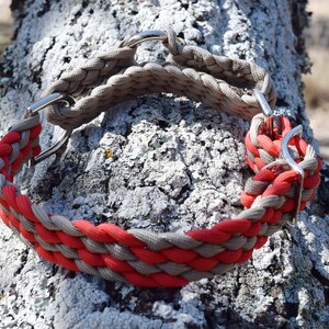 Puede incluir: Un collar trenzado para perros con paracord rojo y gris. El collar presenta herrajes met&aacute;licos plateados y se muestra contra un fondo de tronco de &aacute;rbol texturizado. Dise&ntilde;ado para perros medianos a grandes.