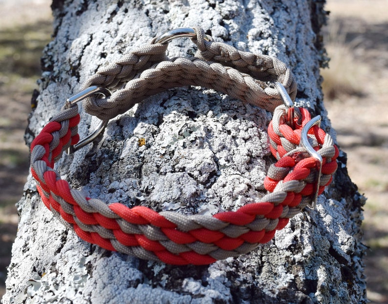 Puede incluir: Un collar trenzado para perros con paracord rojo y gris. El collar presenta herrajes met&aacute;licos plateados y se muestra contra un fondo de tronco de &aacute;rbol texturizado. Dise&ntilde;ado para perros medianos a grandes.