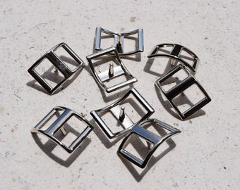 May include: A set of eight silver-toned metal buckles, each with a rectangular shape and a curved design. The buckles are arranged in a random pattern on a light gray surface.