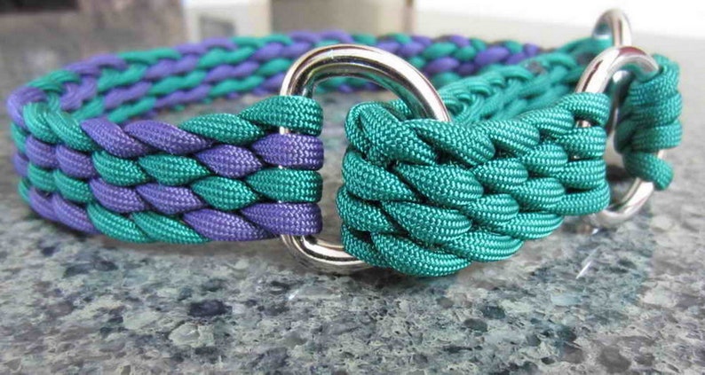 A braided Martingale adjustable dog collar with a silver metal buckle is lying on a gray stone surface. The collar is made of two colors of 650 paracord, one is a teal green and the other is a purple. The collar is braided in a pattern that alternates the two colors.