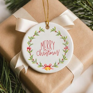 May include: A round white ceramic ornament featuring a hand-painted green mistletoe wreath with red berries and small red bows. The text Merry Christmas is written in red script in the center, with two small yellow bells at the bottom, hanging by a gold cord.