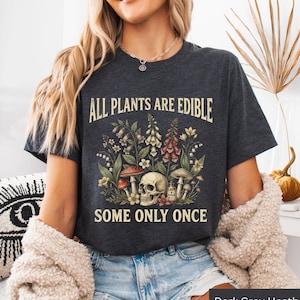 Könnte beinhalten: Ein dunkelgrau meliertes T-Shirt mit einem Grafikdruck, der einen menschlichen Schädel, verschiedene Pilze und Wildblumen zeigt. Der Text "ALL PLANTS ARE EDIBLE" steht darüber und "SOME ONLY ONCE" ist unter dem Schädel und den Pflanzen gedruckt.