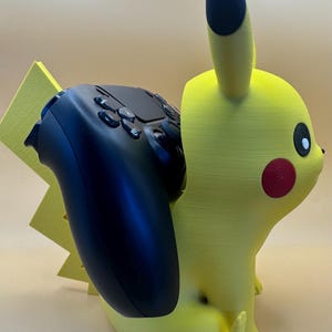 May include: A yellow Pikachu-shaped controller holder with a black controller. The holder has a red cheek and black eyes. The controller is black with multiple buttons. The holder is designed to hold a game controller.