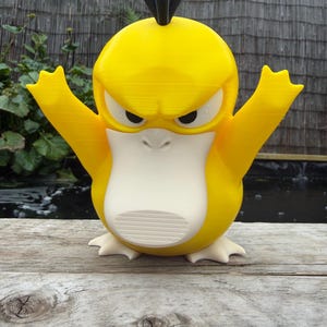 May include: A yellow and white plastic figure of a creature with a large, flat beak, three black prongs on its head, and raised arms. The figure has a stern facial expression with narrow eyes and a wide, pale beak, standing on two small feet.