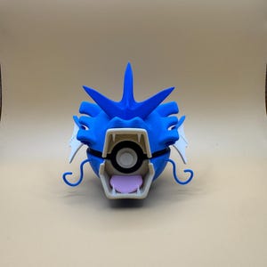 May include: A 3D-printed Gyarados figure in blue and white, a Pokémon character. The figure has a detailed design, with a blue head, white accents, a white mouth, and a pink tongue. The figure is set against a neutral background.