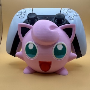 May include: A pink 3D-printed Jigglypuff figure with green eyes and a red mouth, in front of a white and black game controller. The figure has a round body, small arms, and pointed ears. The controller has black joysticks and buttons.