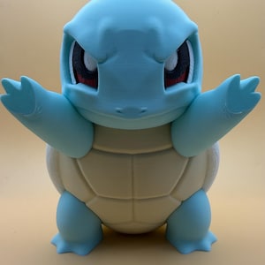 May include: A light blue and cream-coloured figurine of a bipedal turtle creature with a rounded head, large eyes with dark pupils, and outstretched arms. The figure features a smooth, matte finish with a segmented shell on its front torso.